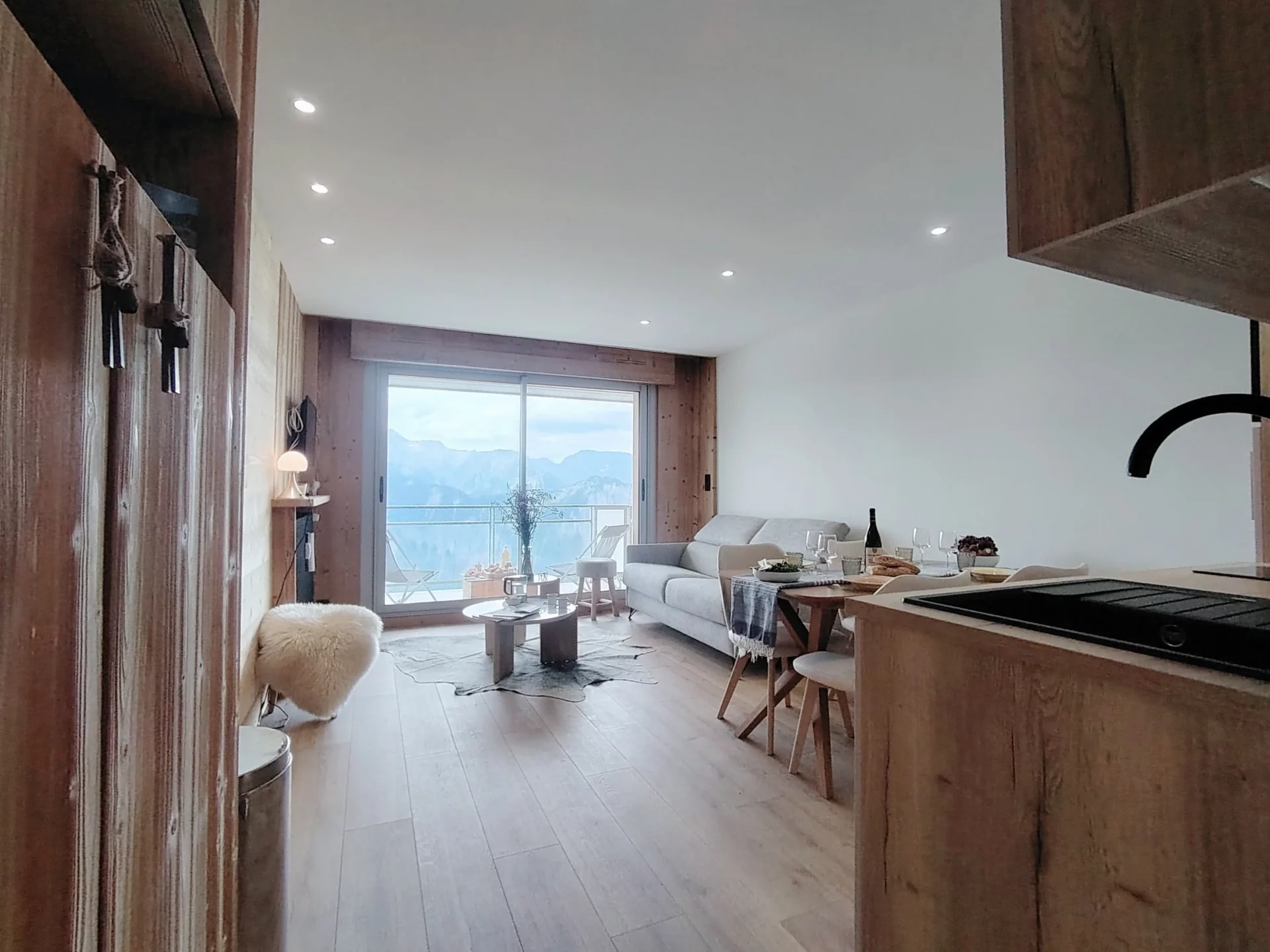 Location appartement à l'alpe d'huez - Pièce de vie avec cuisine au premier plan, table de repas dressée et salon à l'arrière plan avec canapé et tables basses