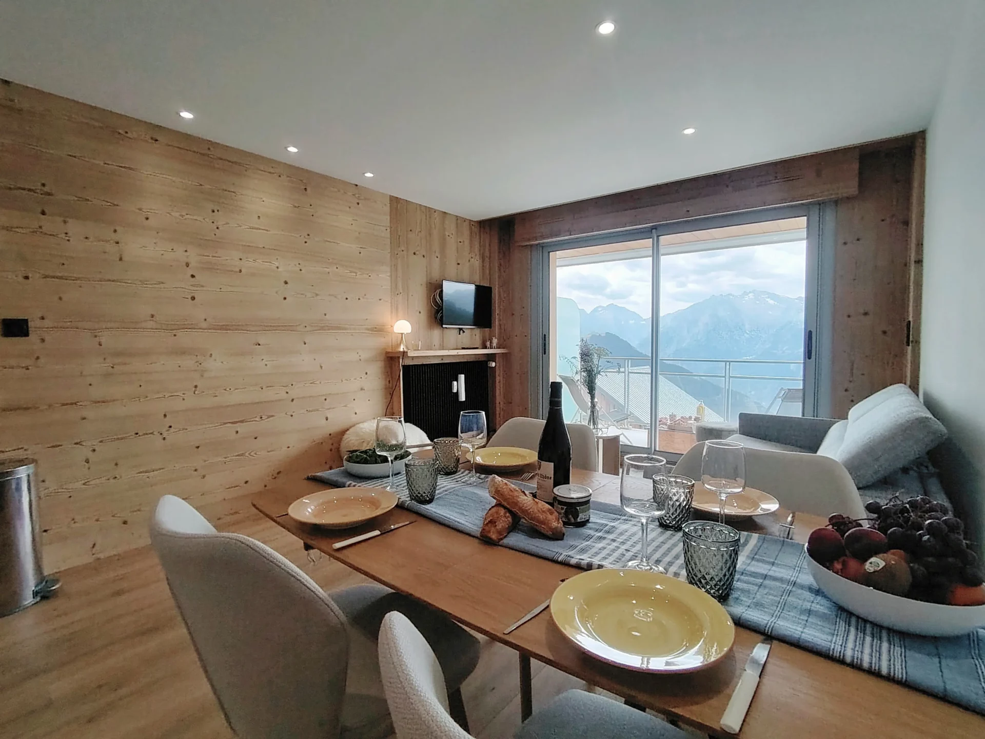 Location appartement à l'alpe d'huez - Pièce de vie avec salon et belle vue sur les massifs. Table dressée au premier plan avec des fruits et des baguettes