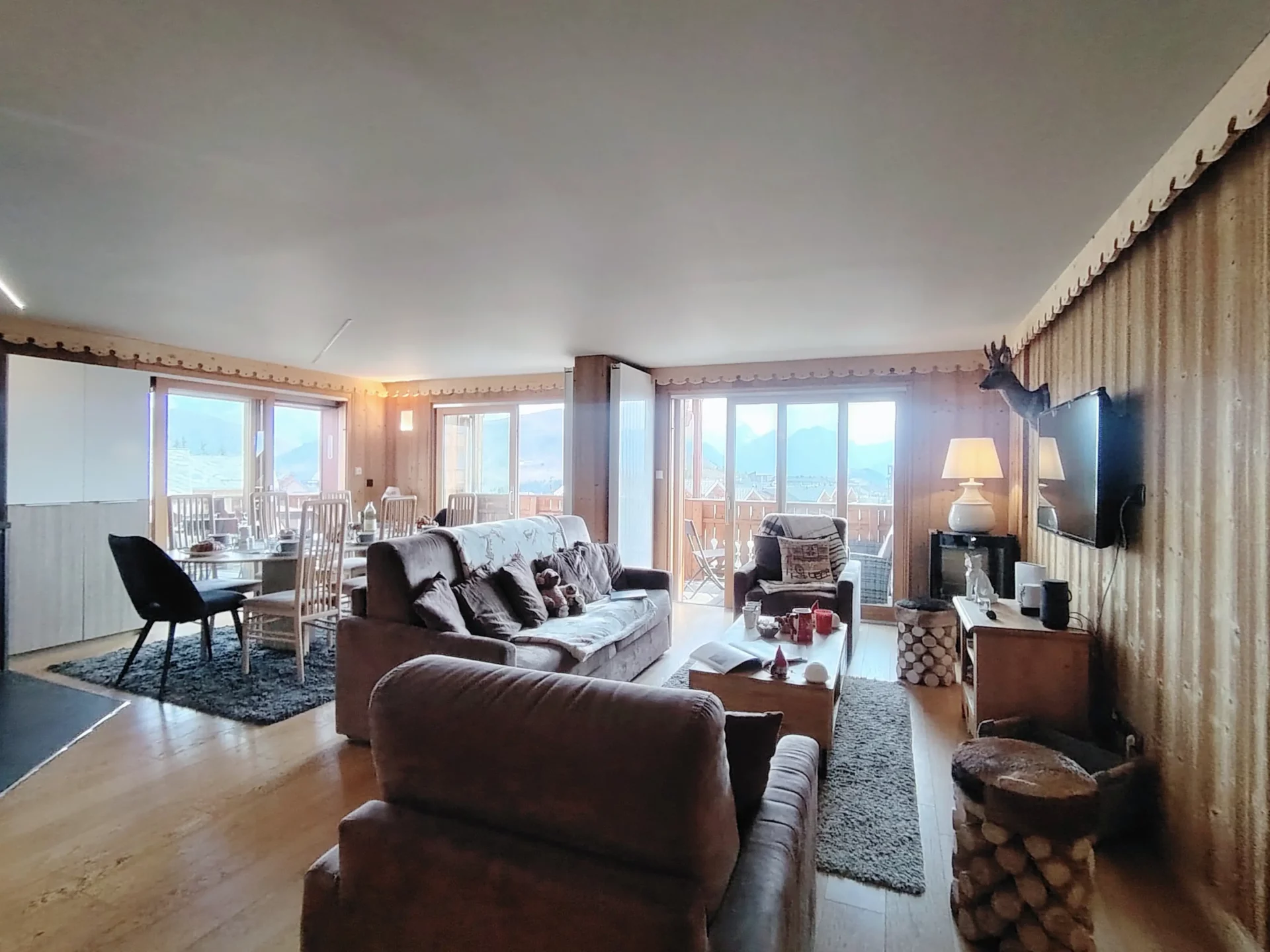 Grand salon lumineux de l’appartement Les Alpages à l’Alpe d’Huez, avec coin repas, baies vitrées et vue panoramique sur les montagnes.