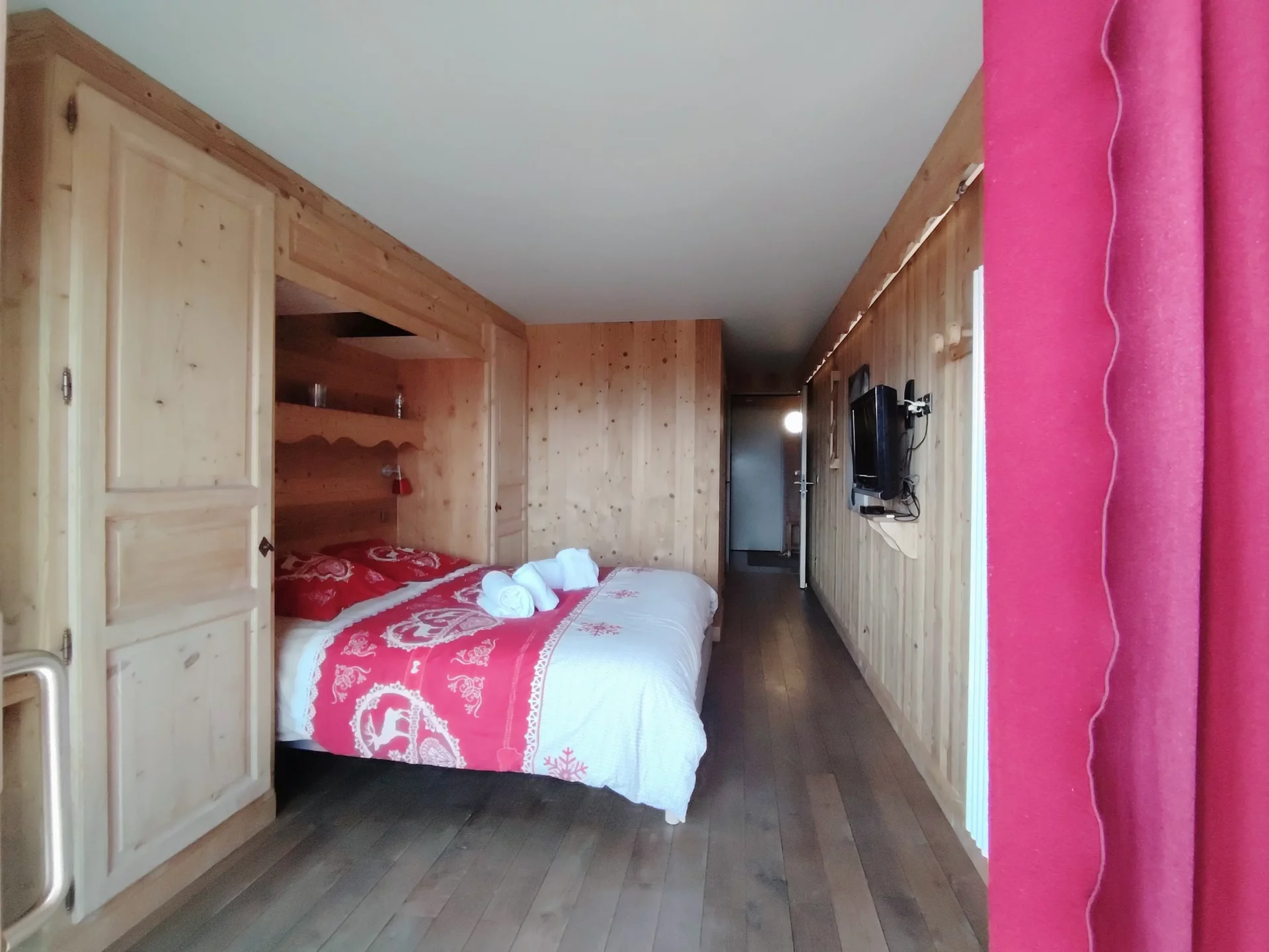 Chambre lumineuse de l’appartement Les Alpages à l’Alpe d’Huez, décor bois et rideaux rouges, avec accès direct au balcon privé