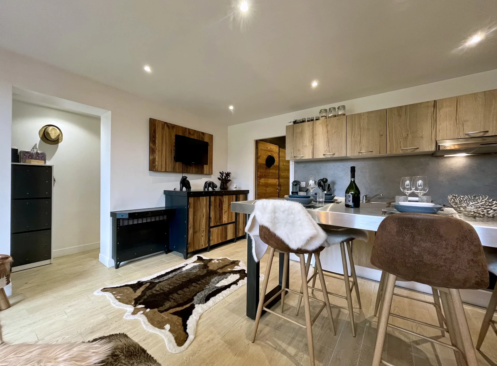 Vue générale du séjour du Chalet Isba à l’Alpe d’Huez, avec cuisine ouverte, coin repas et salon décoré dans un style montagnard moderne avec bois clair et peau de bête.