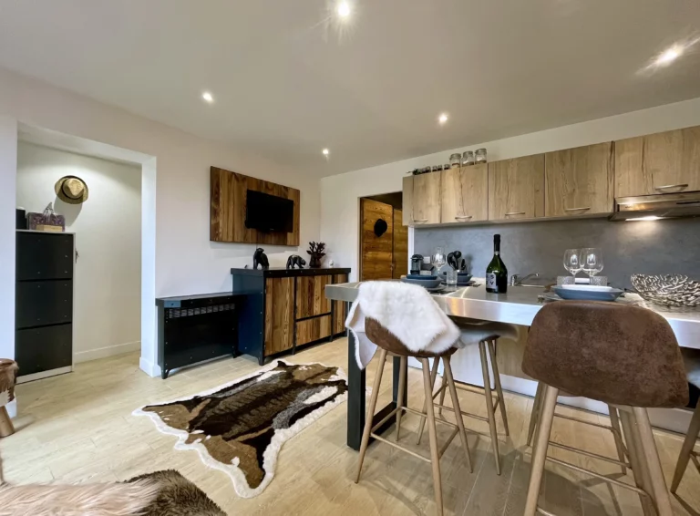 Vue générale du séjour du Chalet Isba à l’Alpe d’Huez, avec cuisine ouverte, coin repas et salon décoré dans un style montagnard moderne avec bois clair et peau de bête.