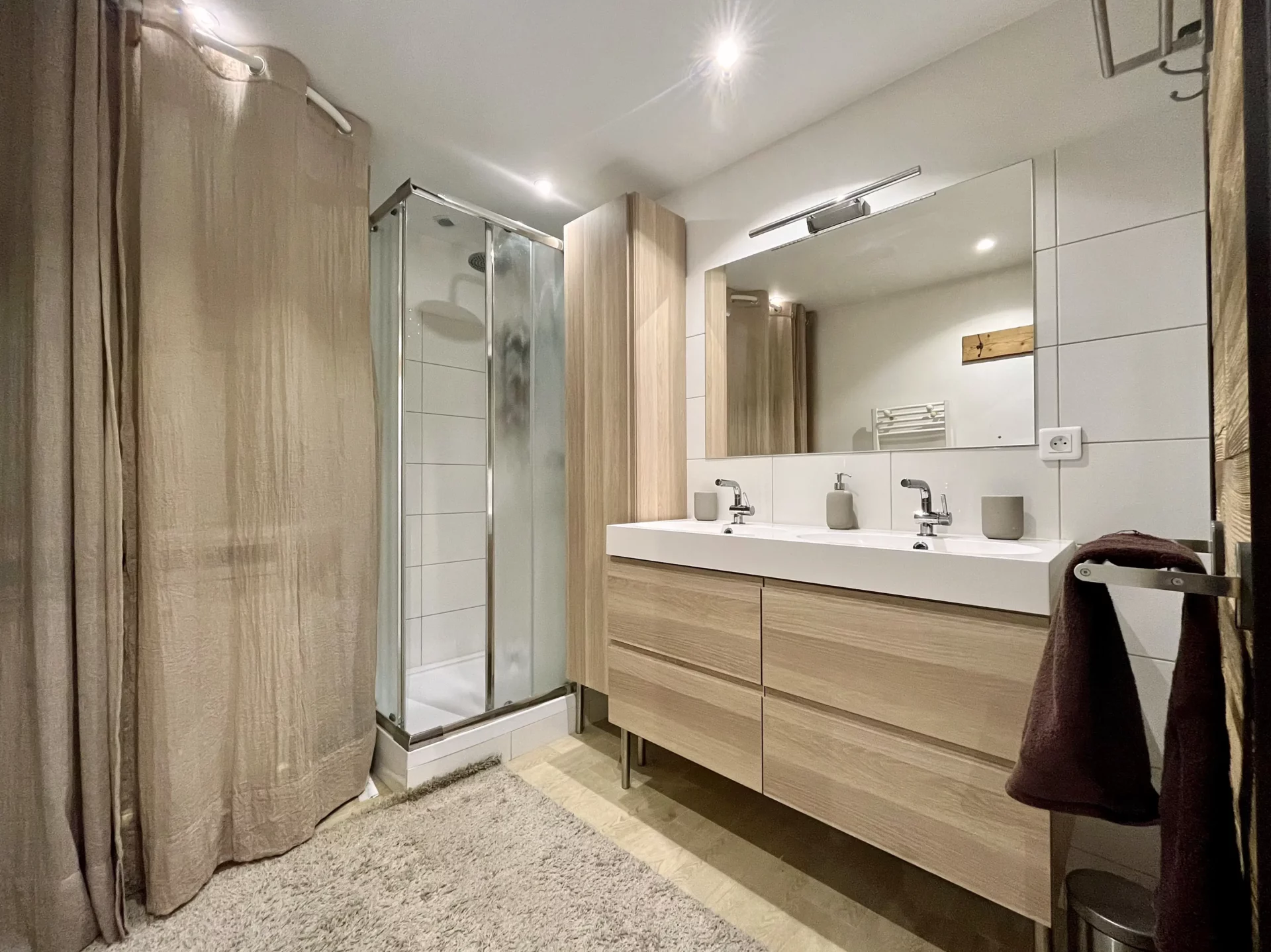 Salle de bain du Chalet Isba à l’Alpe d’Huez avec douche vitrée, meuble double vasque en bois clair et coin buanderie équipé d’un lave-vaisselle dissimulé derrière un rideau beige.