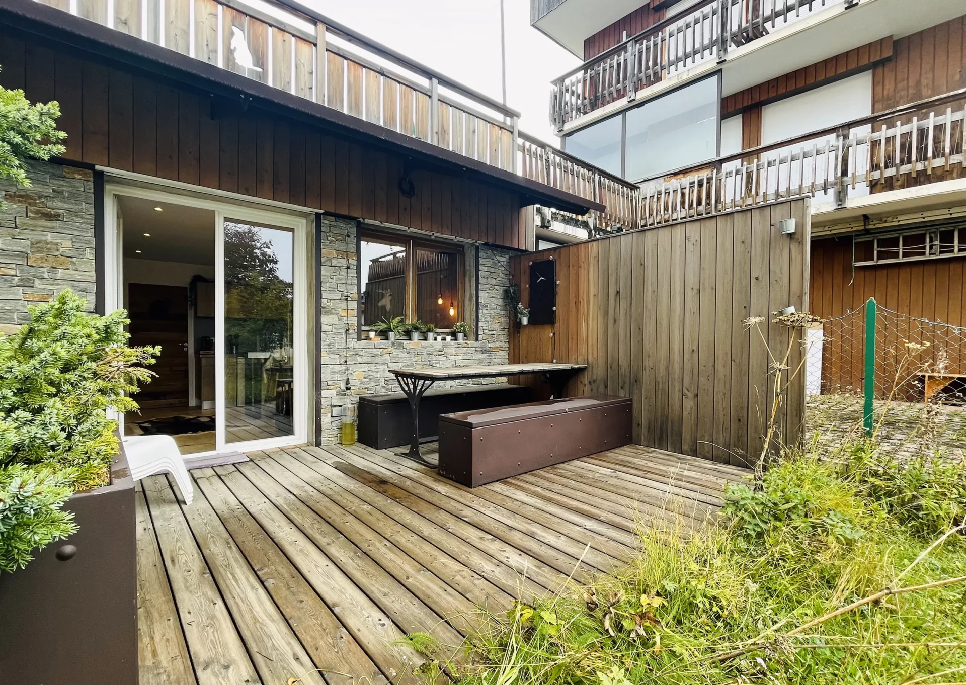 Façade extérieure du Chalet Isba à l’Alpe d’Huez, terrasse aménagée en bois avec bancs et table, mur en pierre et accès direct au jardin.