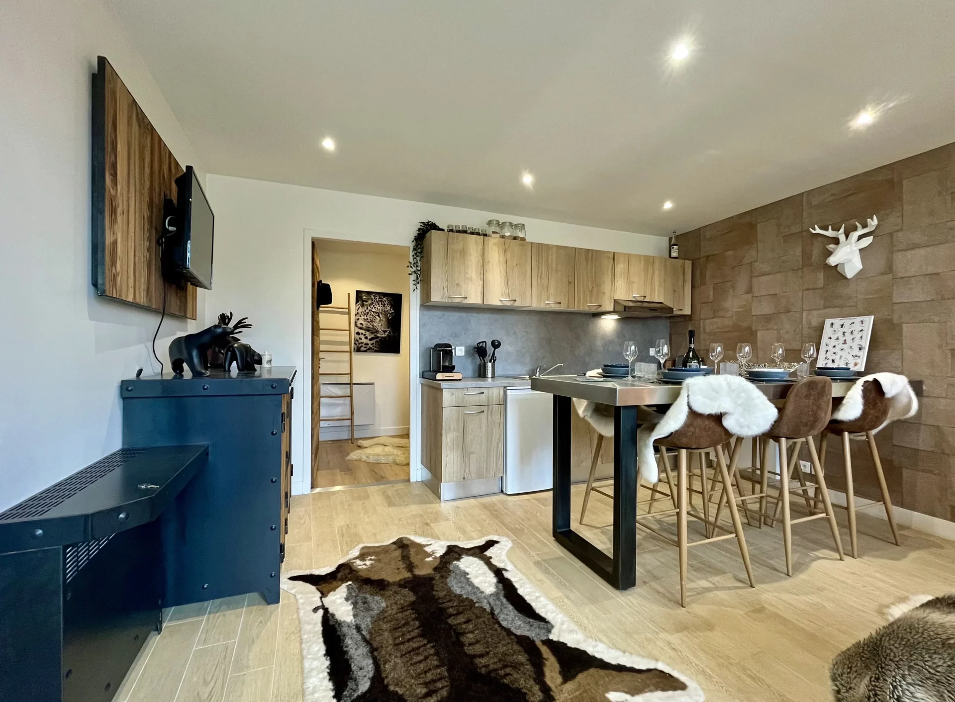 Pièce à vivre lumineuse du Chalet Isba à l’Alpe d’Huez, avec table haute, tabourets en velours, cuisine équipée et décoration murale façon pierre naturelle.