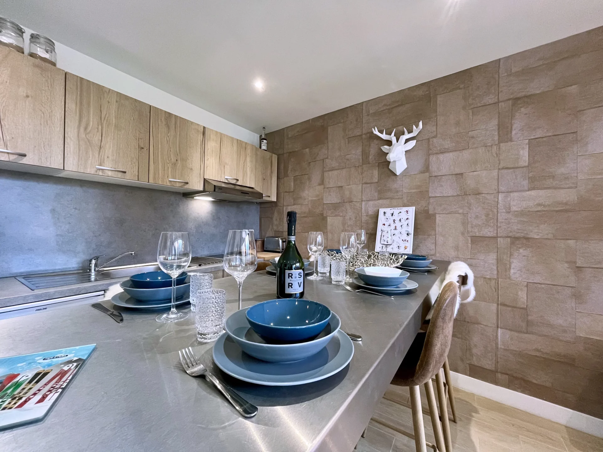 Coin repas du Chalet Isba à l’Alpe d’Huez, table haute en bois et métal, vaisselle bleue et mur effet pierre avec tête de cerf décorative.