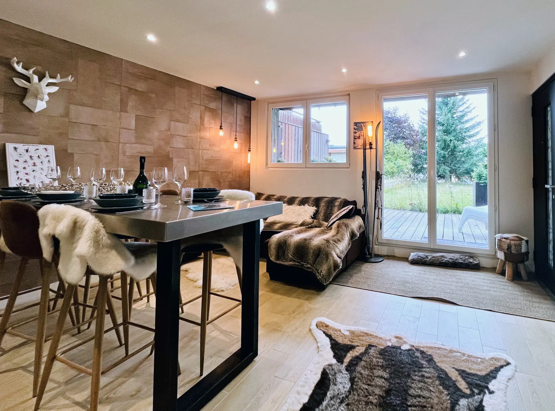 Salon du Chalet Isba avec canapé d’angle en fausse fourrure, éclairage tamisé et grande baie vitrée donnant sur la terrasse et le jardin.