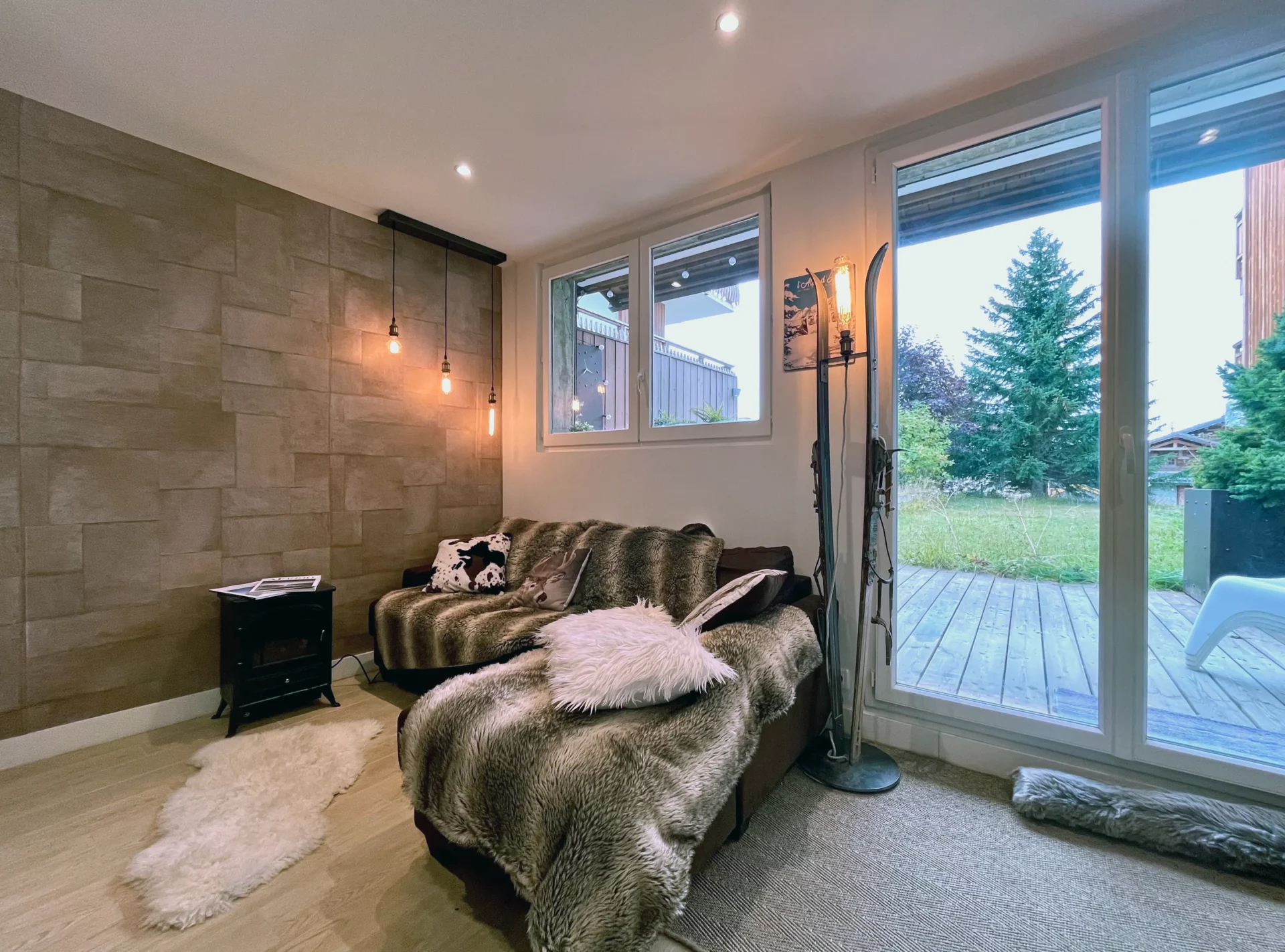 Coin salon chaleureux du Chalet Isba à l’Alpe d’Huez, avec canapé couvert de plaids, lampe sur pied et vue sur la terrasse en bois et les sapins.