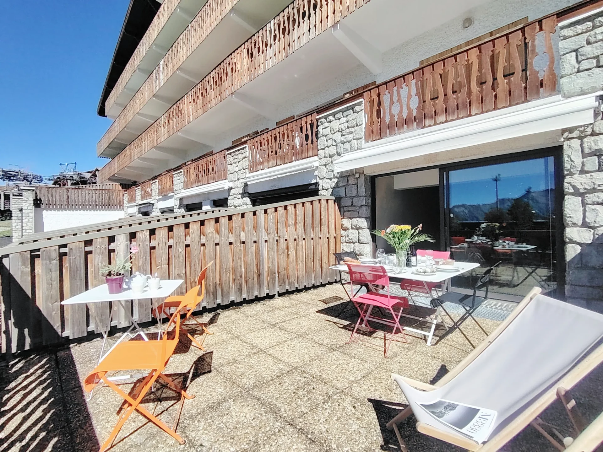 Grande terrasse ensoleillée de l’appartement à louer à l’Alpe d’Huez, équipée de tables et chaises colorées