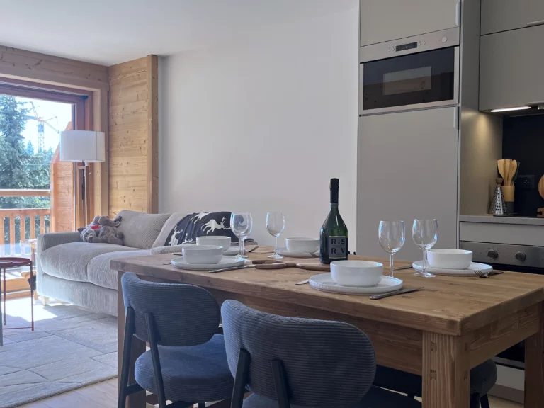 Séjour lumineux avec canapé beige, table en bois dressée et cuisine moderne équipée dans un appartement à l’Alpe d’Huez.