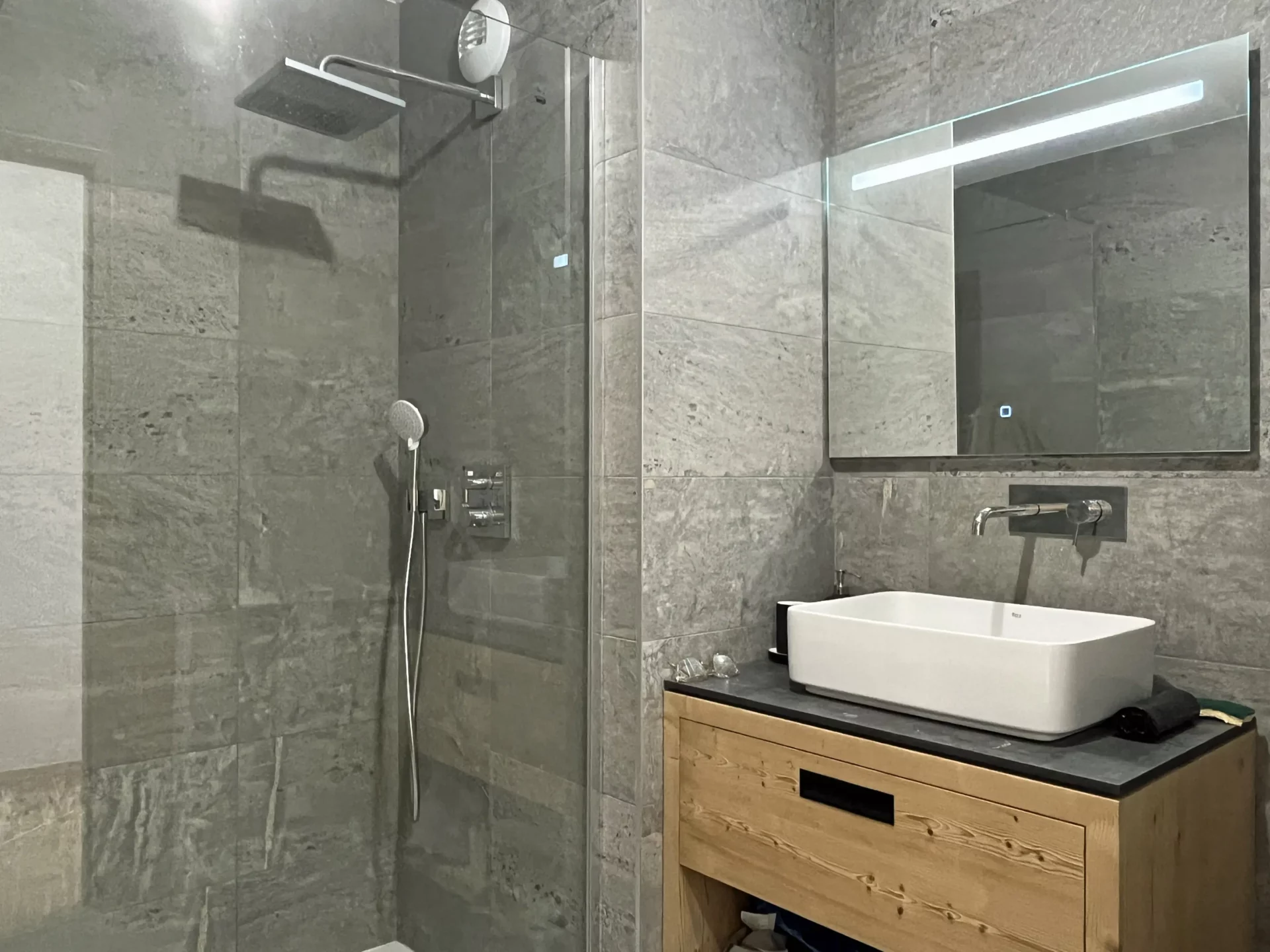 Salle de bain design avec douche à l’italienne, vasque blanche sur meuble en bois et mur en pierre grise.