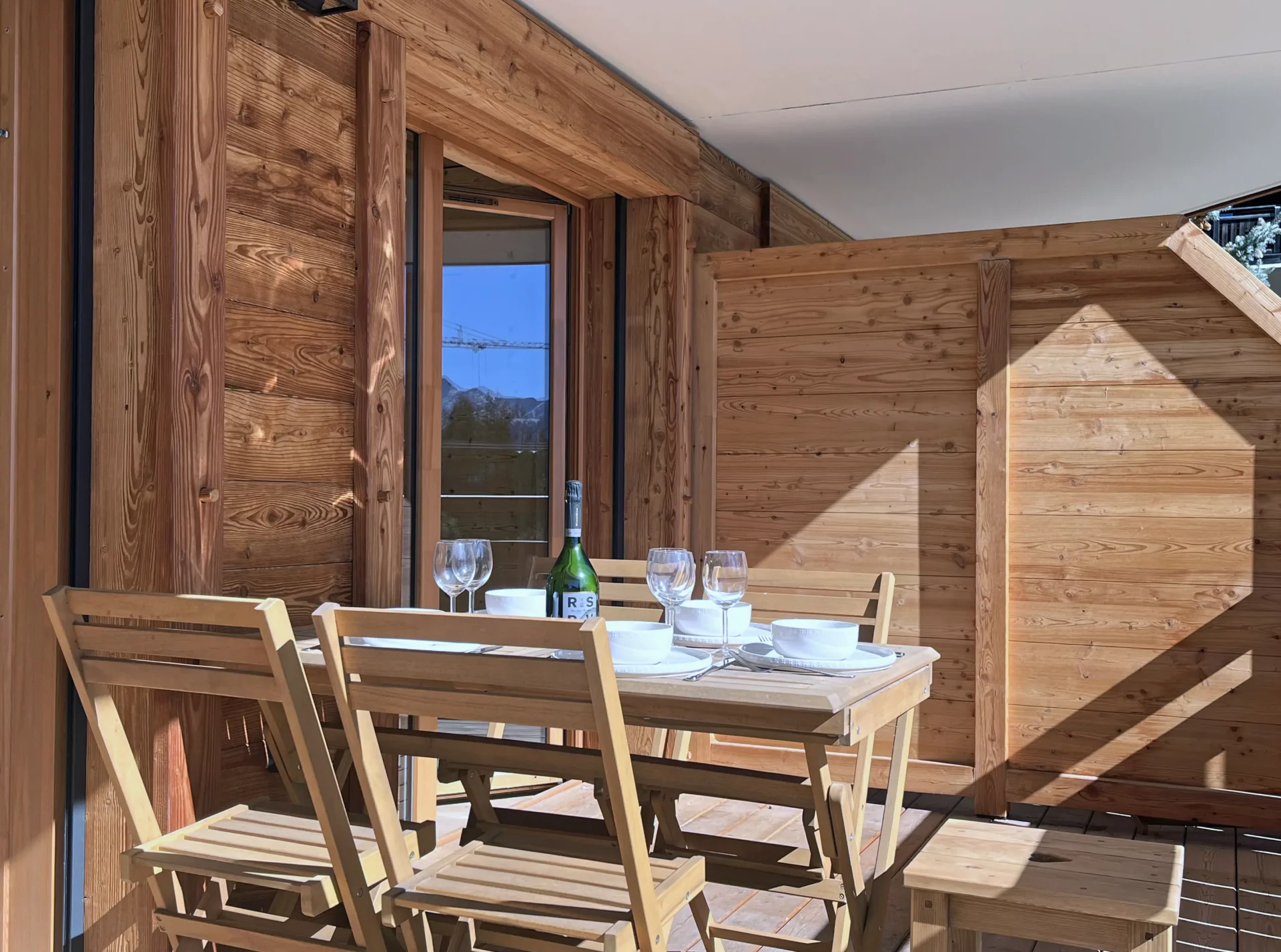 Terrasse en bois avec table dressée, quatre chaises et bouteille de vin, exposée au soleil dans la résidence Cachemire à l’Alpe d’Huez.