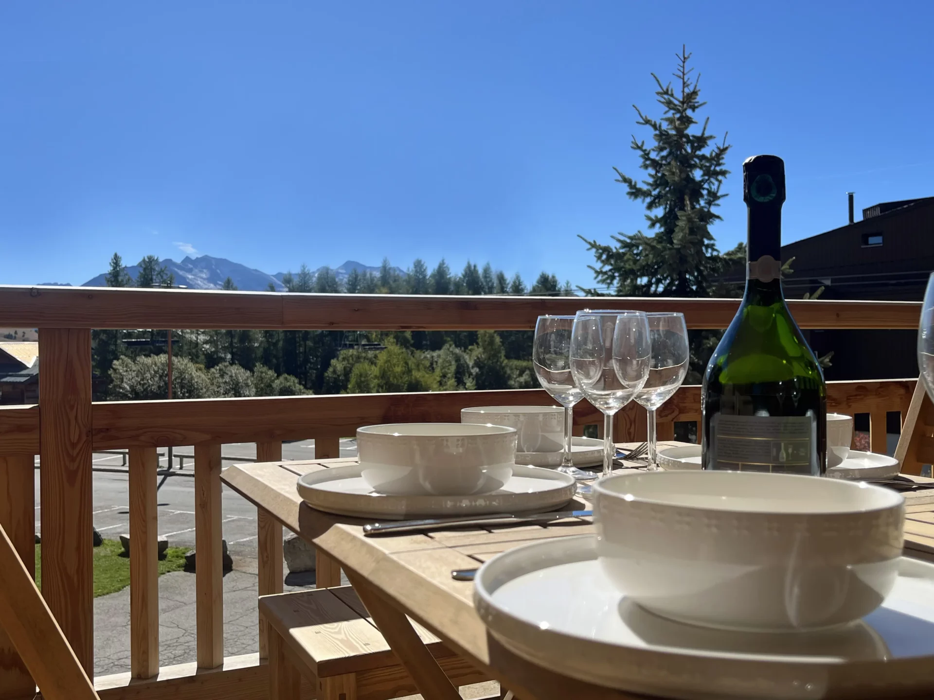 Gros plan sur la table dressée en terrasse avec verres et assiettes, face aux montagnes sous un ciel bleu.