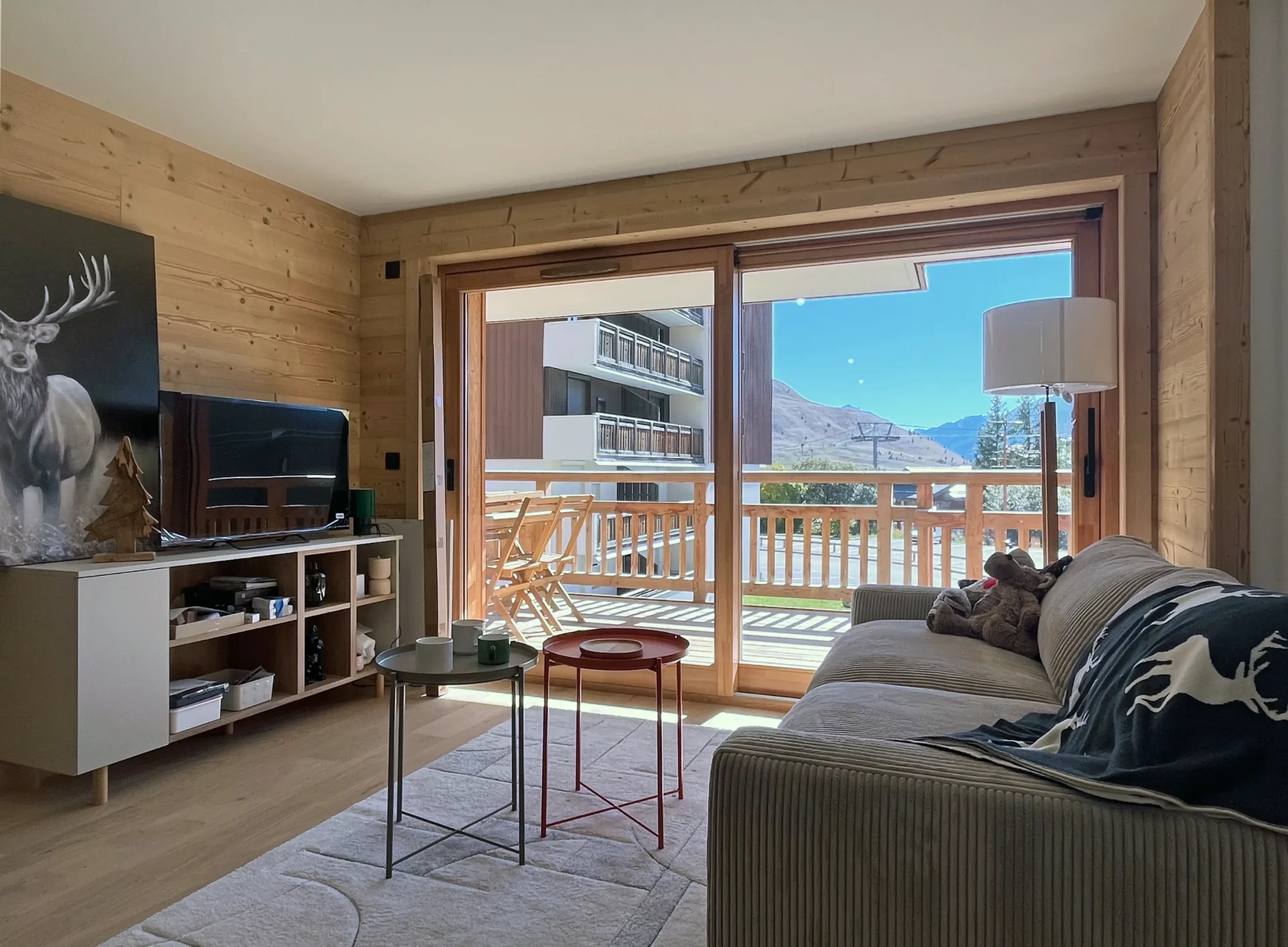 Salon chaleureux avec grande baie vitrée donnant sur le balcon ensoleillé et vue sur les montagnes de l’Alpe d’Huez.