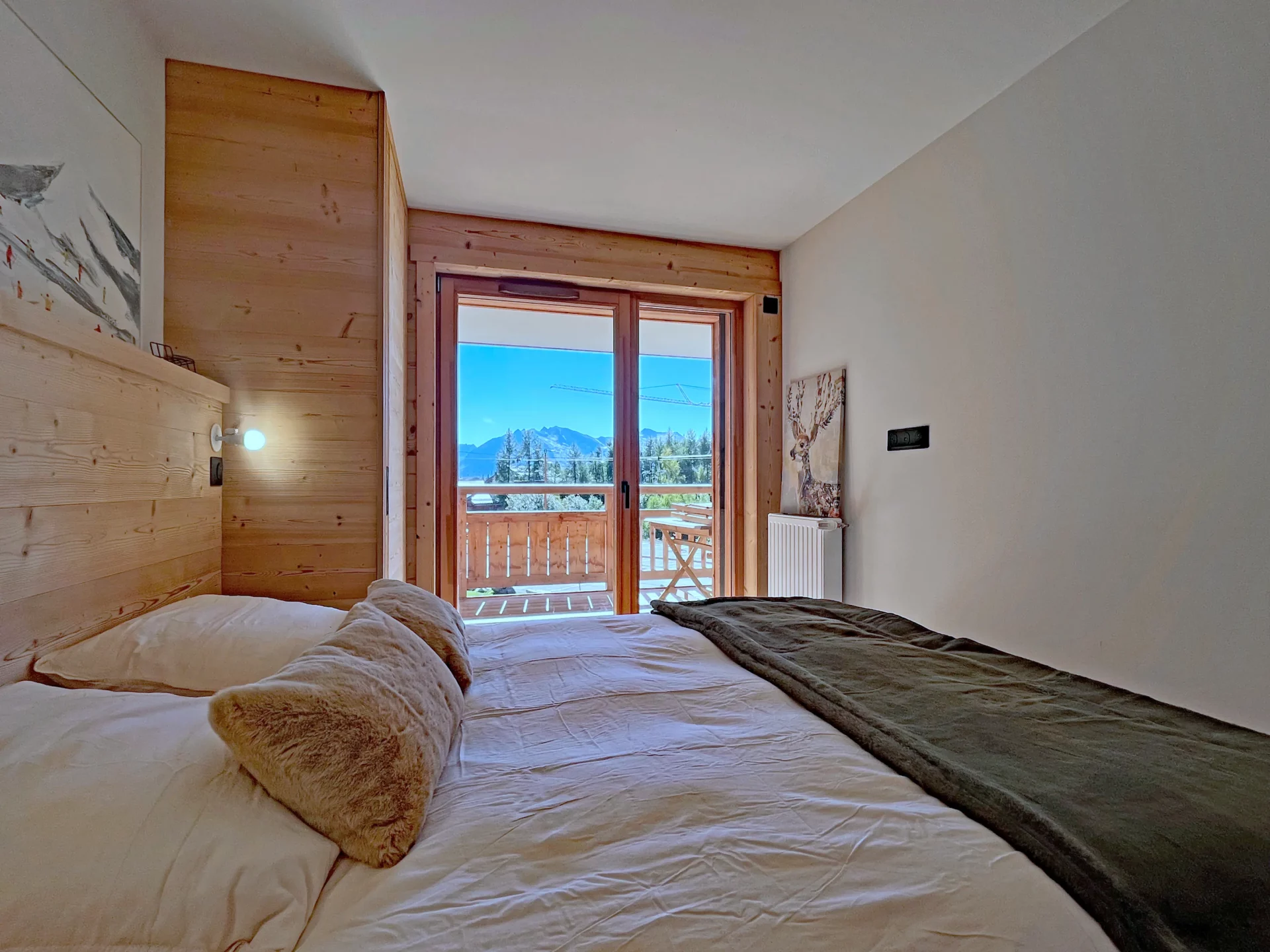 Chambre lumineuse avec lit double, plaid vert et balcon offrant une belle vue sur les montagnes de l’Alpe d’Huez.