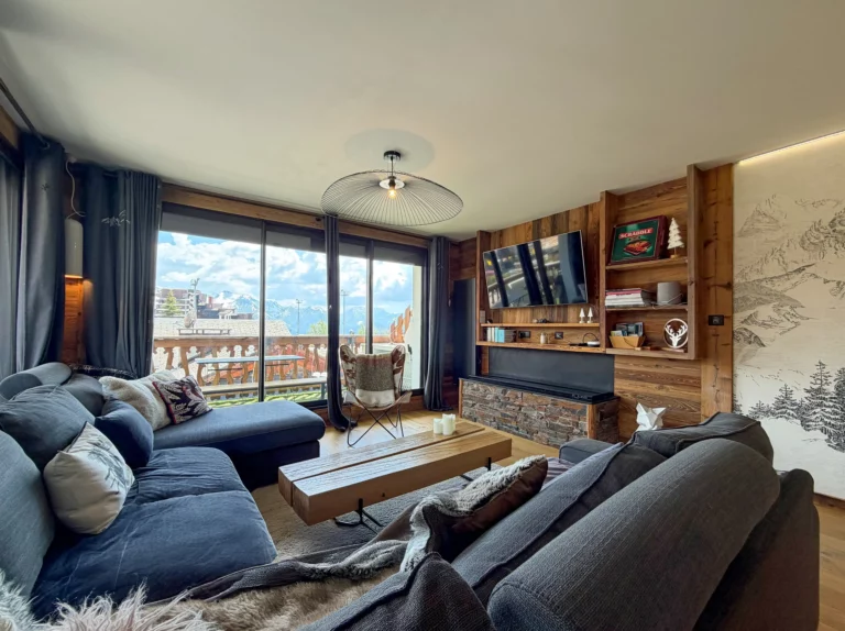 Vaste pièce de vie pouvant accueillir 9 personnes avec un canapé d'angle faisant face à un mur décoratif avec télévision et cheminée à vapeur. Les baies vitrées laissent voir le balcon sur lequel une table vous permet de prendre vos repas tout en contemplant la vue sur la piscine et les montagnes.
