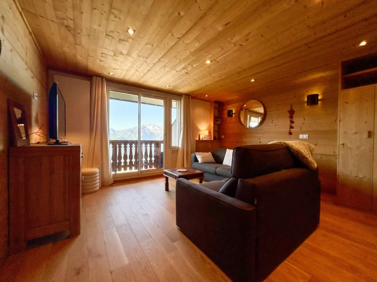 Pièce de vie tout en bois avec canapé devant la télévision et les baies vitrées ouvrant sur les sommets montagneux.