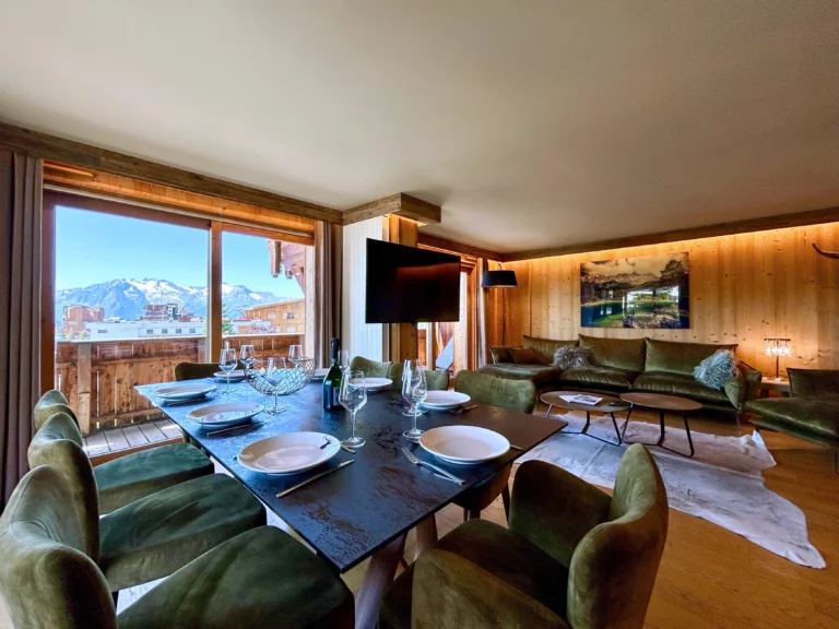 Belle table de repas en decton, dressée pour 8 personnes devant de larges baies vitrées ouvrant sur une vue panoramique. Une télévision centrale permet de se distraire pendant que vous êtes assis sur les canapés en velours gris dans l'arrière plan