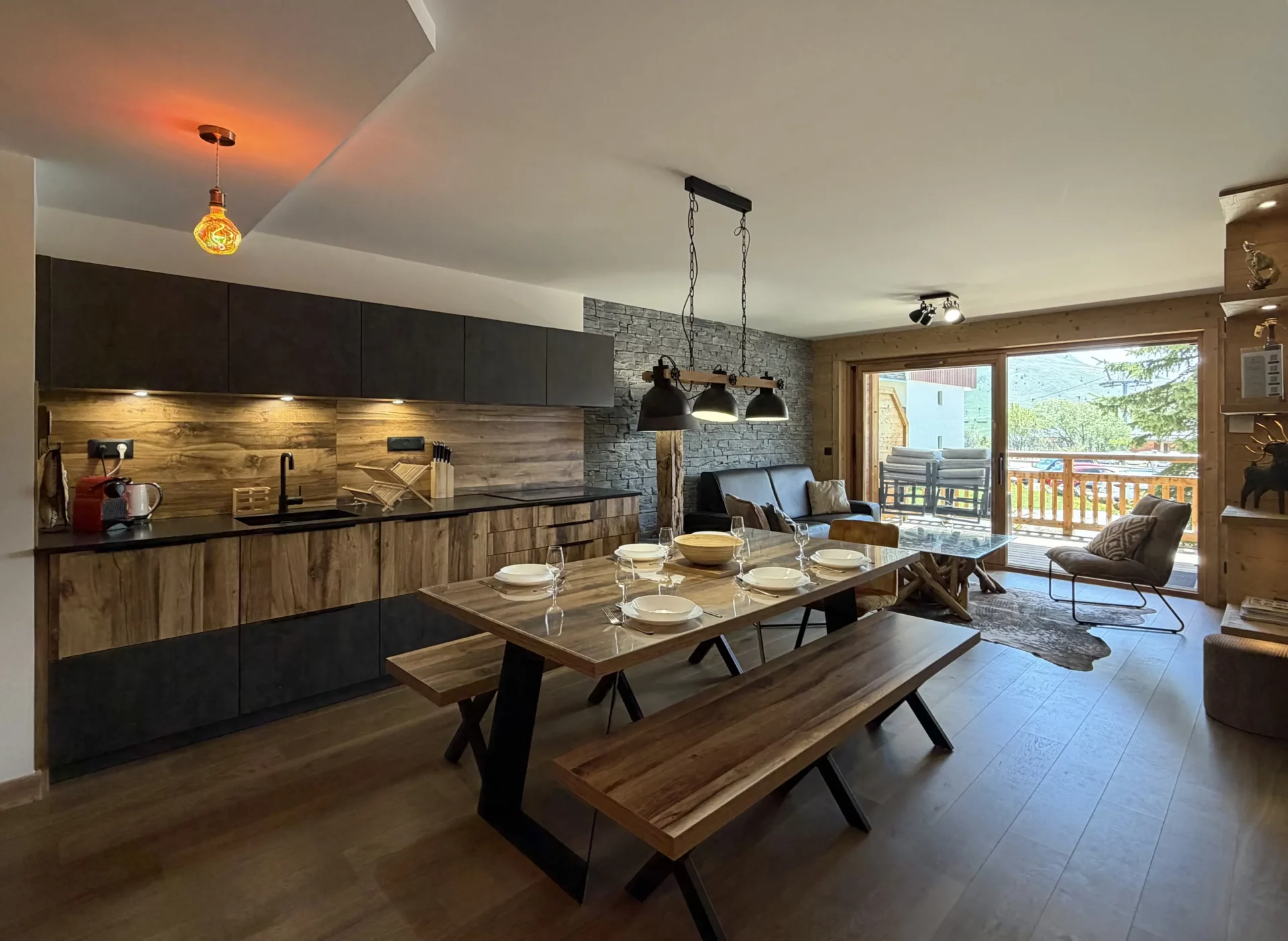 Grande table de repas avec des bancs comme assise devant une grande cuisine entièrement équipée. A l'arrière plan, un canapé permet de regarder la télévision ou de contempler la vue par les fenêtres ouvrant sur les massifs montagneux.