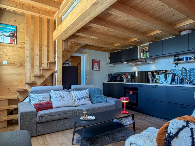 Pièce de vie cozy avec le salon équipé d'un canapé devant la table basse, d'une cuisine équipée sur la droite et d'un escalier en arrière plan, permettant d'accéder à la chambre en mezzanine.